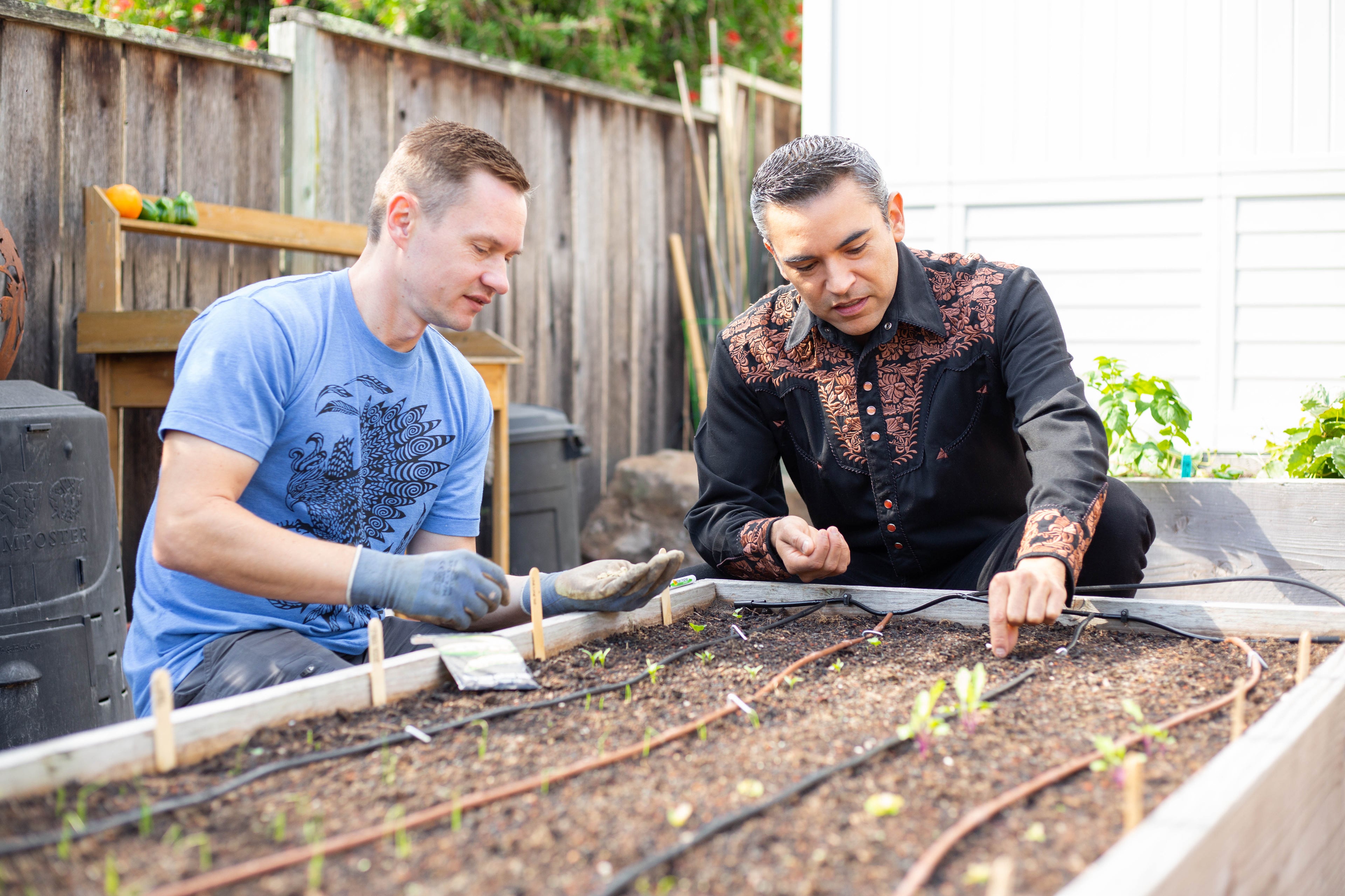 Two men working together in a garden, one wearing a blue shirt and the other in a black patterned shirt.